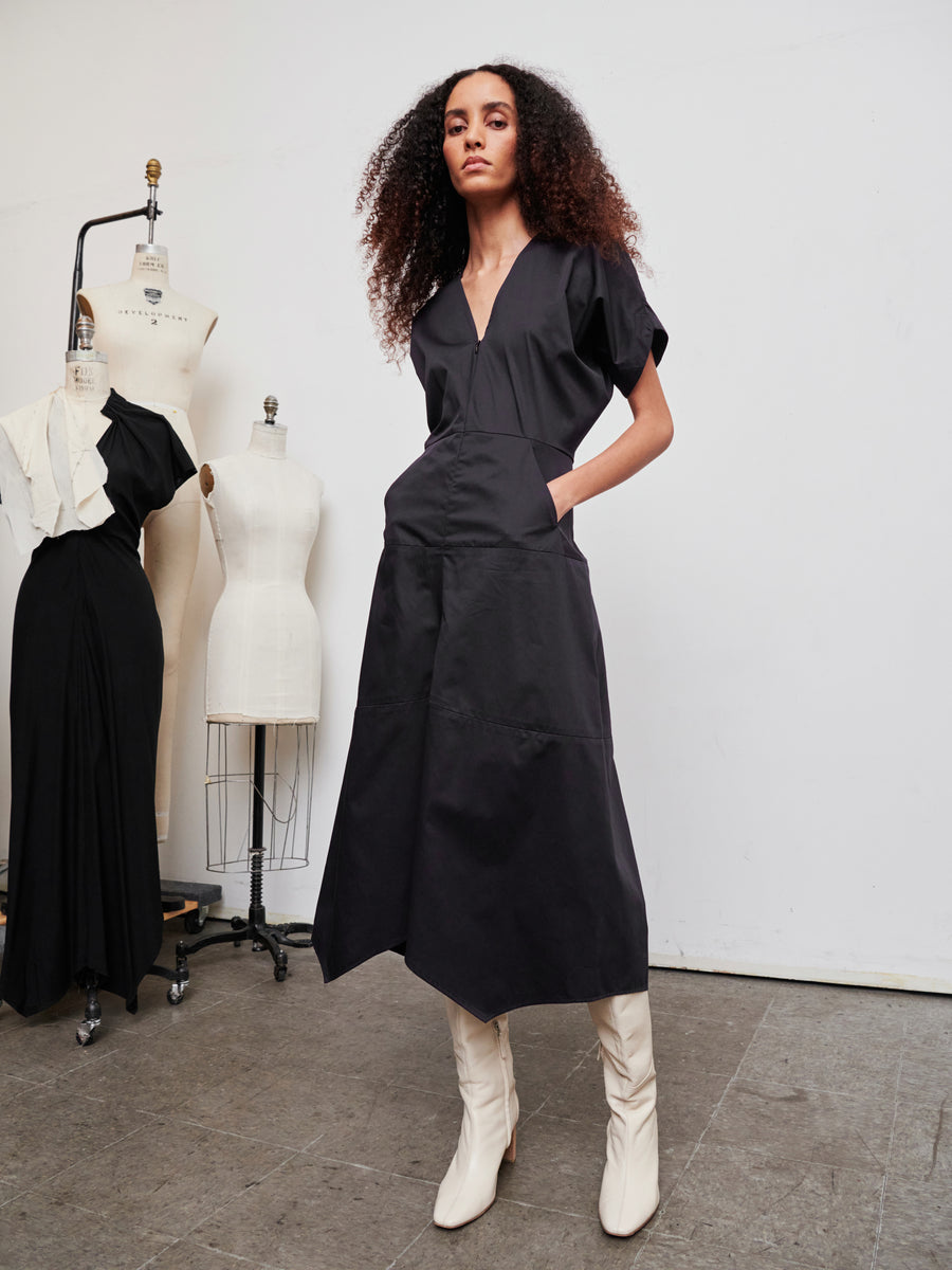 A person with curly hair stands confidently in a black dress and white boots in a fashion studio. In the background, two dress forms are positioned against a plain white wall, one of which is displaying the elegant Aissa Joi Dress by Zero + Maria Cornejo, adorned with ruffles.