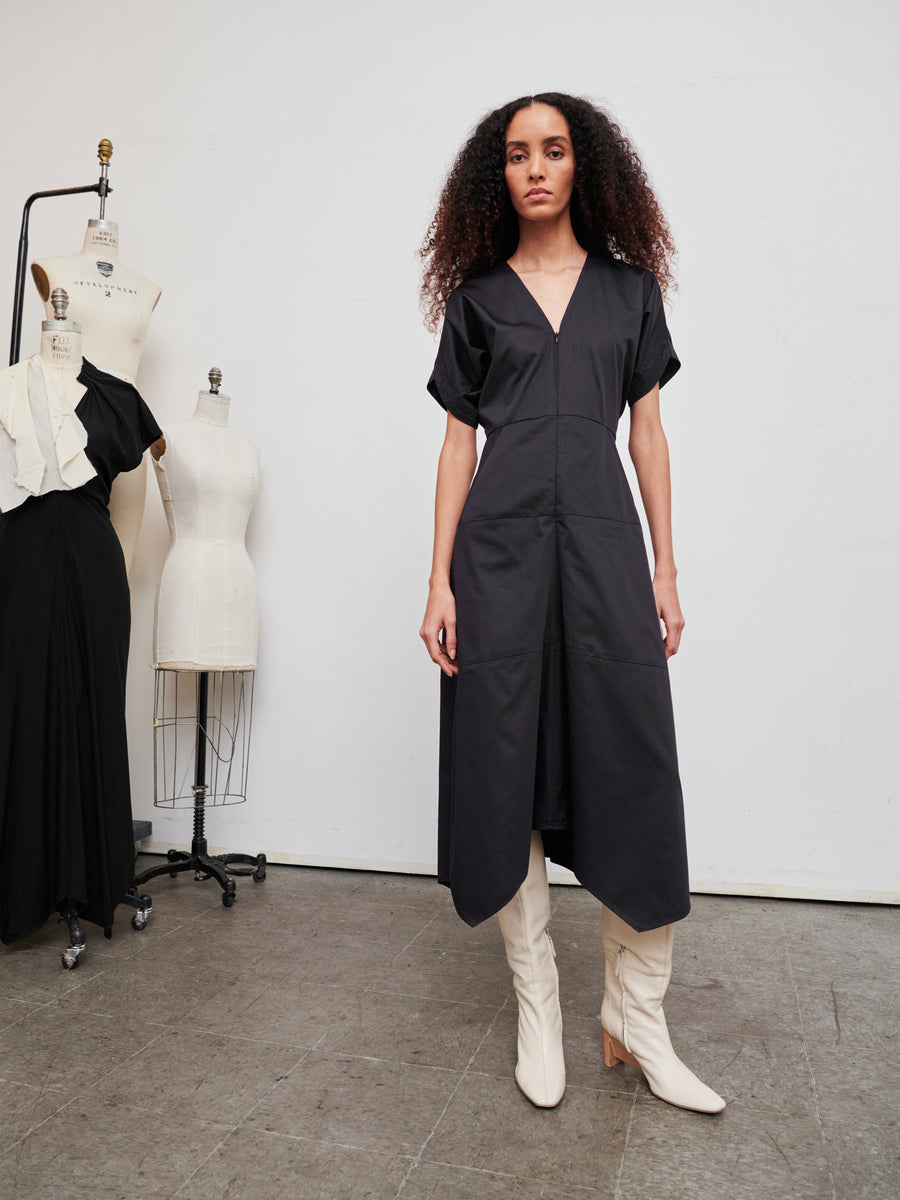 In a fashion studio, a woman with long, curly hair stands elegantly, dressed in the sleek Aissa Joi Dress by Zero + Maria Cornejo and wearing white knee-high boots. Mannequins draped in black and white fabric are positioned against the plain white wall, accentuating the contemporary ambiance of the space.