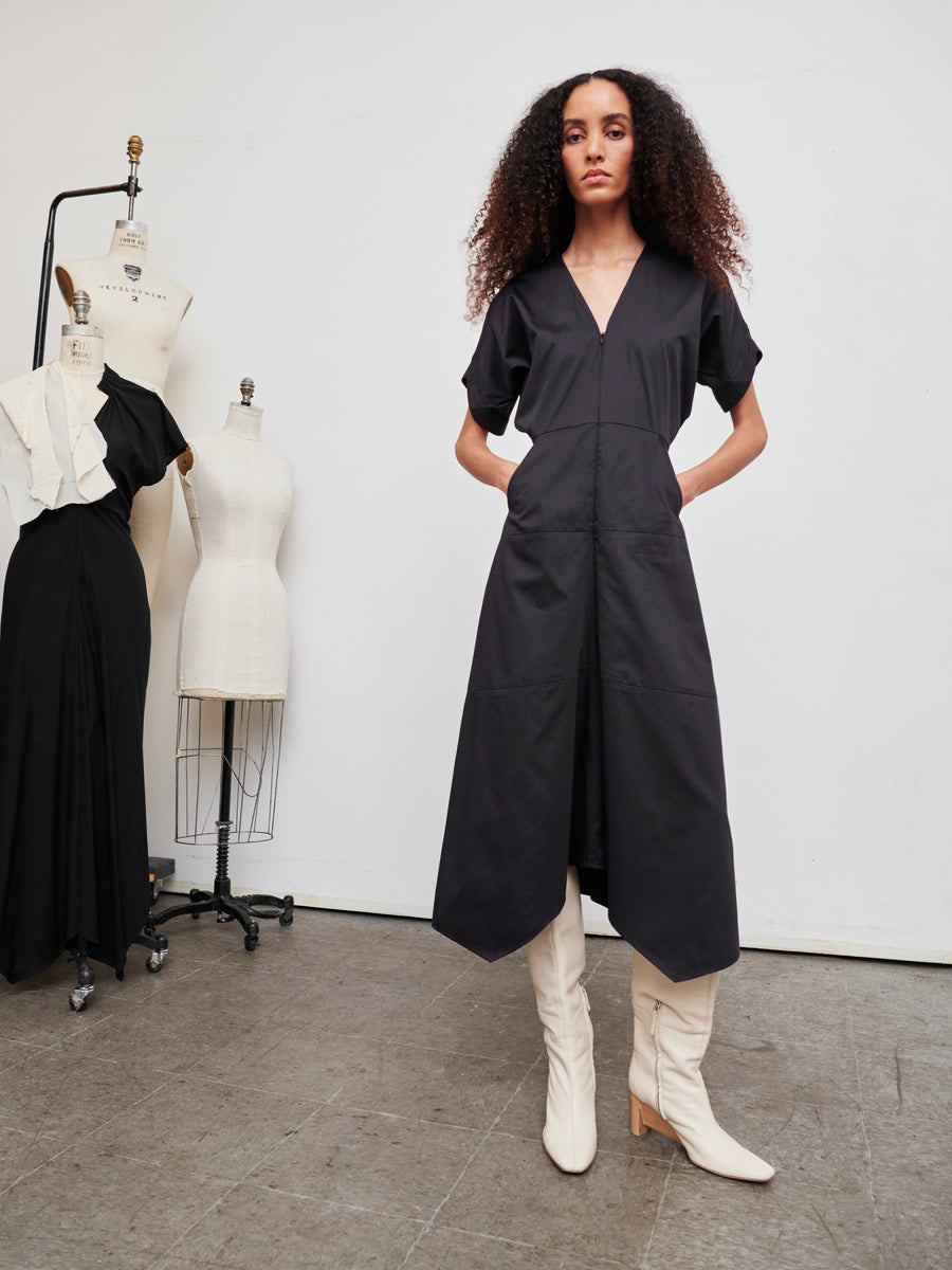 A woman with curly hair stands confidently in a black cotton broadcloth dress and white knee-high boots. She is surrounded by dress forms, one of which features the elegant Aissa Joi Dress by Zero + Maria Cornejo, draped gracefully over it in a studio setting.