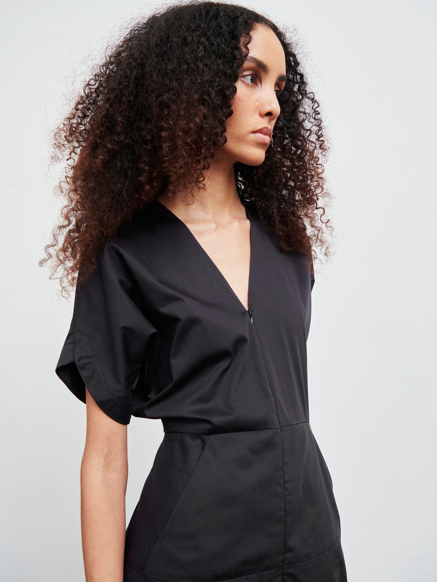 A person with curly hair is wearing the Aissa Joi Dress, a black V-neck broadcloth piece by Zero + Maria Cornejo. They have a serious expression and are looking to the side against a plain, light-colored background.