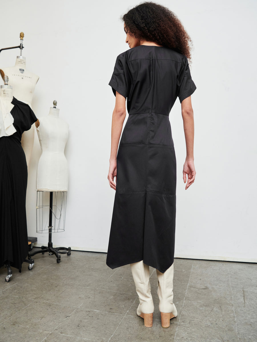 A person with curly hair stands in a studio, showcasing the Aissa Joi Dress by Zero + Maria Cornejo and wearing white knee-high boots. They face away from the camera, surrounded by mannequins on a gray floor.