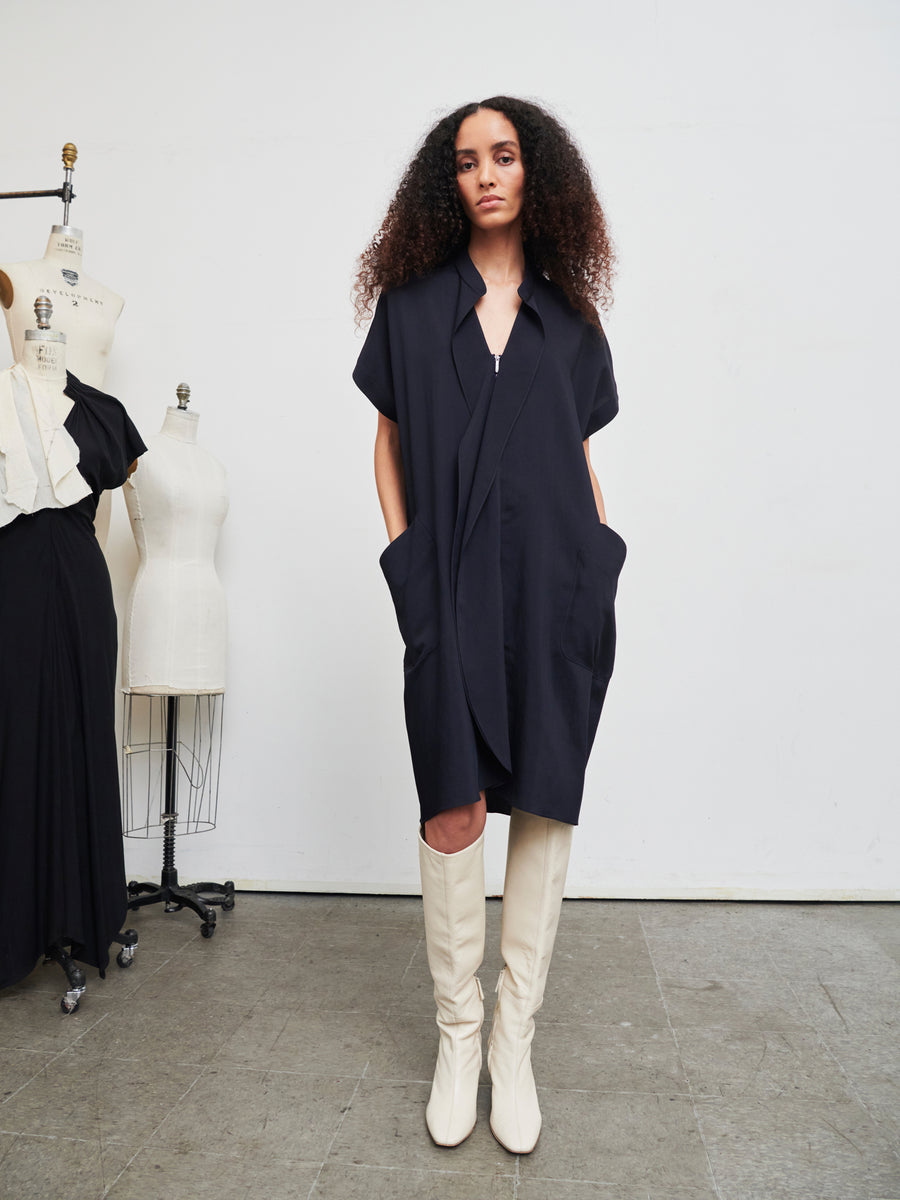 A person with long, curly hair confidently wears the Fin Gaban Dress by Zero + Maria Cornejo, a stylish black dress featuring a draped collar. They pair it with white knee-high boots while standing in a room filled with dress forms and mannequins against a plain white wall, embodying elegance and eco-consciousness in fashion crafted from recycled viscose.