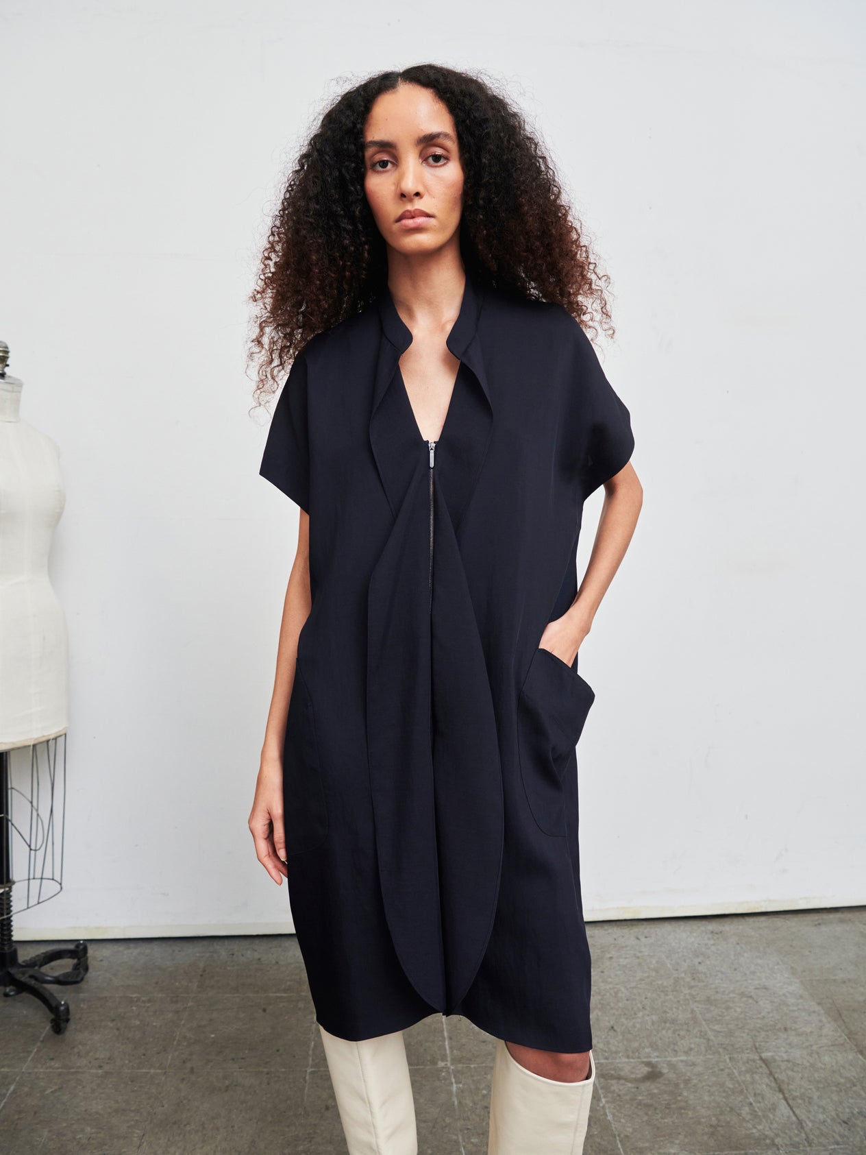 A person with curly hair stands confidently in the Fin Gaban Dress by Zero + Maria Cornejo, an asymmetrical, knee-length dress crafted from recycled viscose, paired with black slip-on shoes. Their hands rest in the dress pockets, while a dress form is visible on the left side against a plain white wall.
