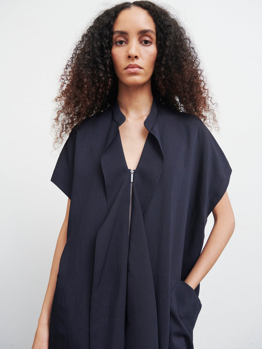 A person with long curly hair wears the Fin Gaban Dress by Zero + Maria Cornejo, a dark knee-length dress featuring a front zipper and pockets. The plain background accentuates the outfit's minimalist and stylish look, crafted from recycled viscose.