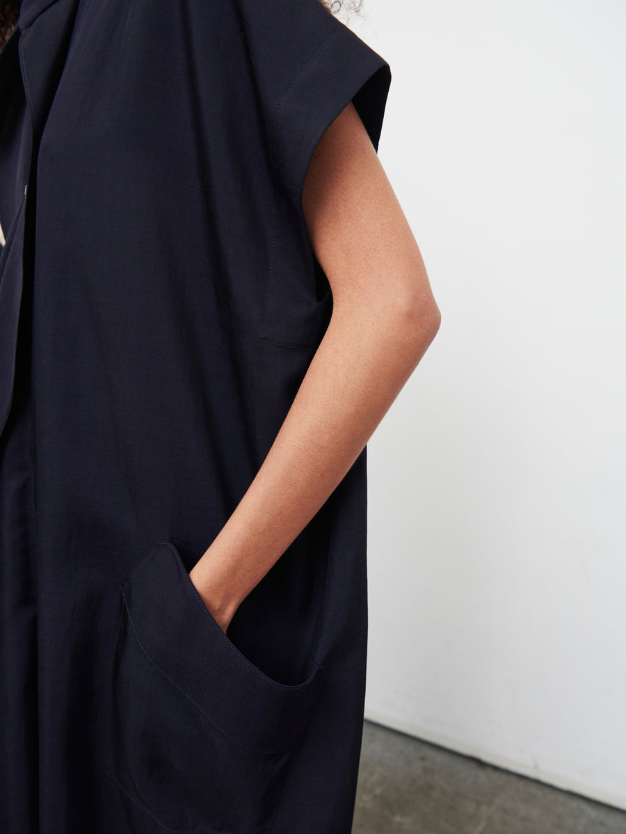 In a minimalist setting, someone stands wearing the Fin Gaban Dress by Zero + Maria Cornejo. This sleeveless black dress features a knee-length hem and a draped collar, with one hand casually slipped into its pocket. The plain white wall and gray floor set off the elegance of this timeless ensemble.