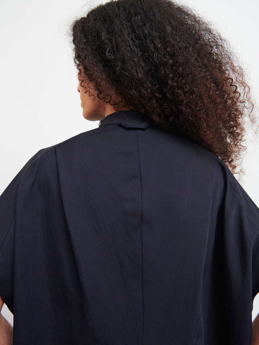 A person with curly hair wearing the Fin Gaban Dress by Zero + Maria Cornejo, seen from behind, stands elegantly. The subtle draped collar of the dress adds sophistication to the look.