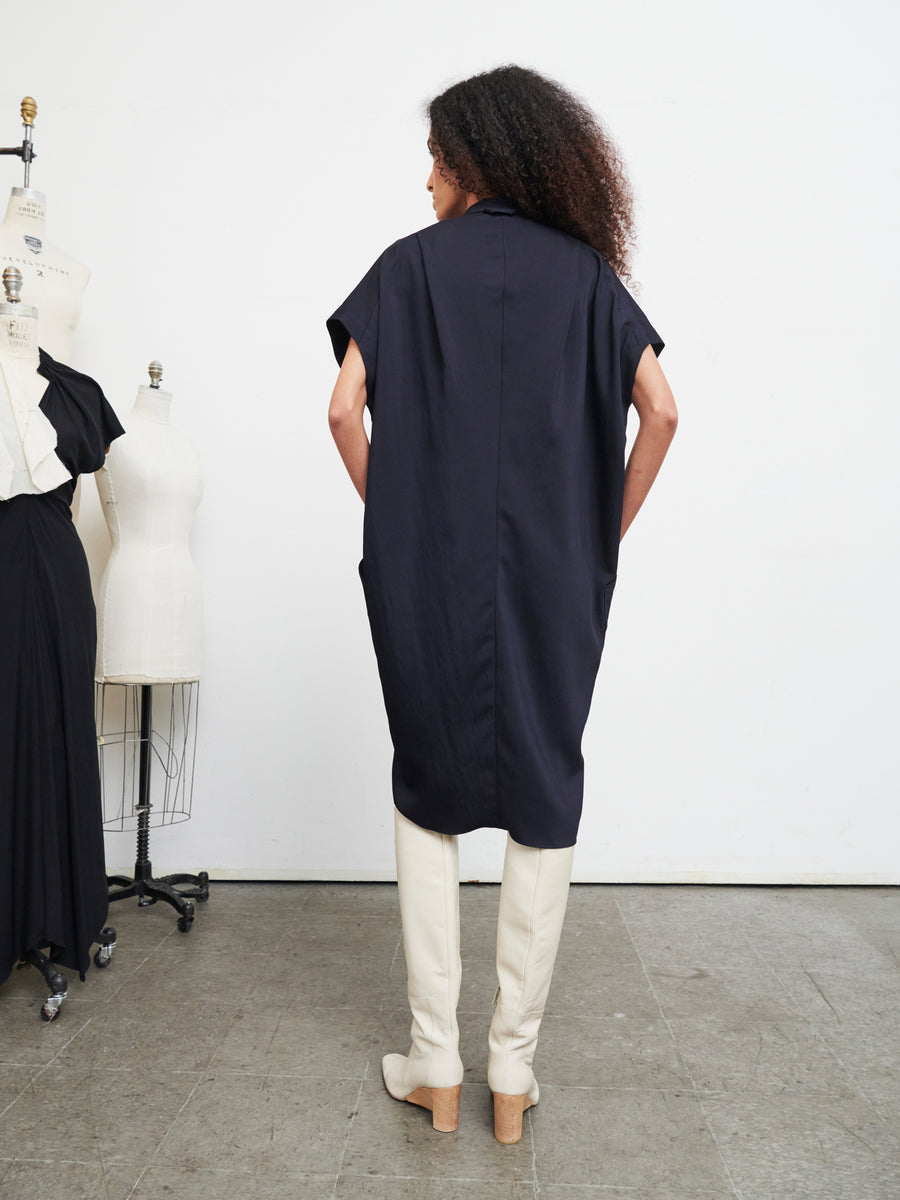 A person with long curly hair stands with their back to the camera, wearing the loose-fitting Fin Gaban Dress from Zero + Maria Cornejo, which features a draped collar and is paired with knee-high white boots. Two dress mannequins are visible in the background.