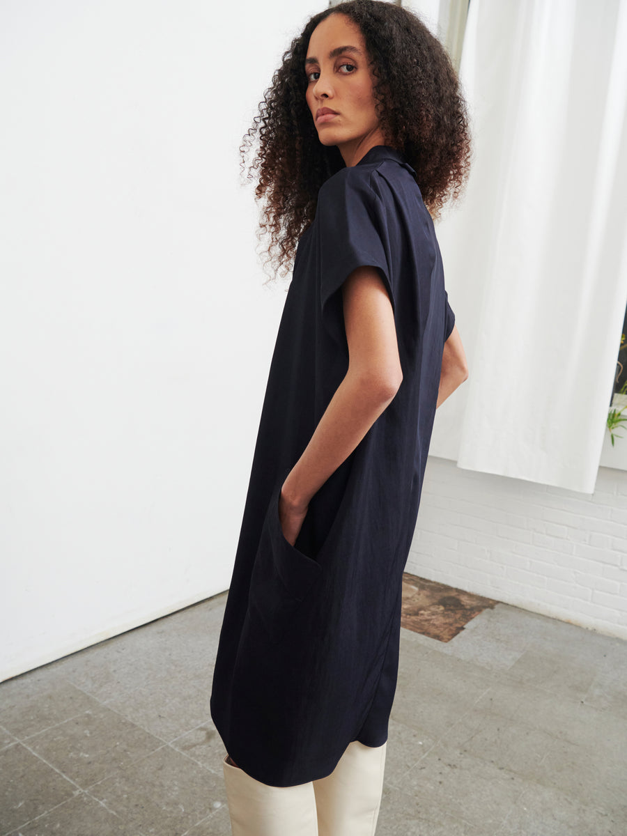 In a minimalist room bathed in natural light, a person with curly hair stands gracefully, showcasing the Fin Gaban Dress by Zero + Maria Cornejo. The navy knee-length dress crafted from recycled viscose is paired stylishly with white pants. They face slightly away from the window adorned with white curtains, one hand resting casually in a pocket.