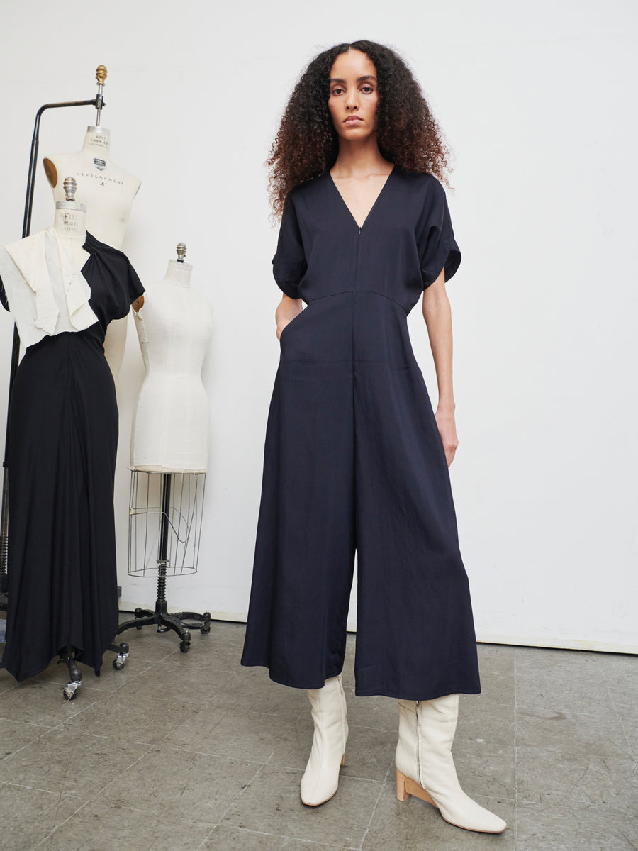 A person with curly hair wears the Aissa Eve Jumpsuit by Zero + Maria Cornejo and white boots, standing in a room with three dress forms. Two of the forms have black and white garments draped over them.