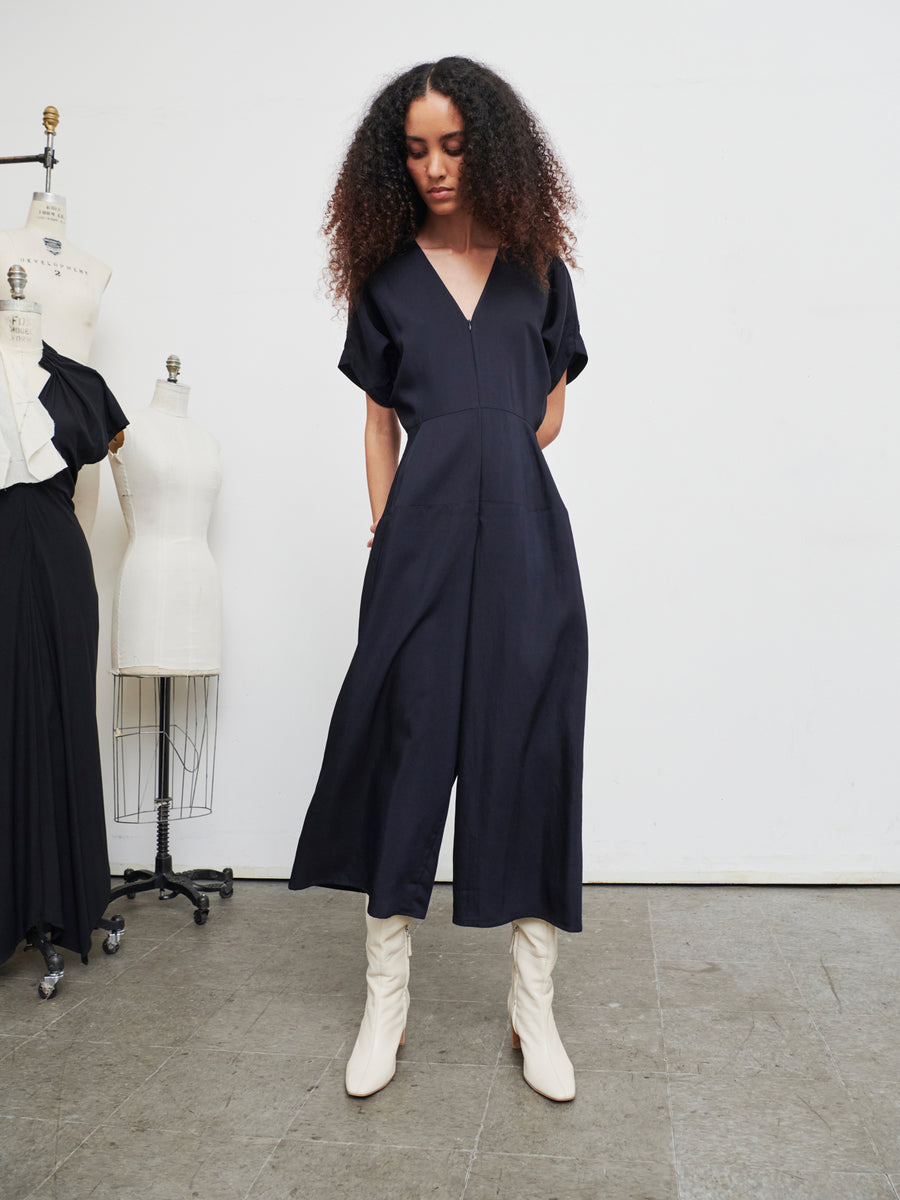 A woman with curly hair stands in a minimalist room wearing the black Aissa Eve Jumpsuit from Zero + Maria Cornejo, paired with white boots. Tailor mannequins with dresses fill the background, highlighting the chic contrast of her outfit's cropped wide leg design.