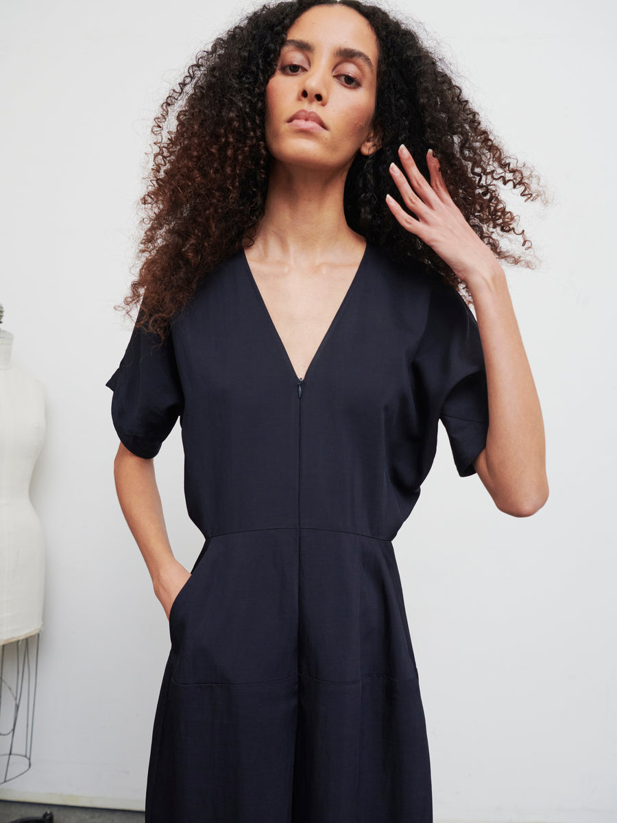 A person with long curly hair is wearing an Aissa Eve Jumpsuit by Zero + Maria Cornejo, gently touching their hair with one hand. They are standing in a minimalist setting with a neutral expression, and a mannequin is partly visible in the background.