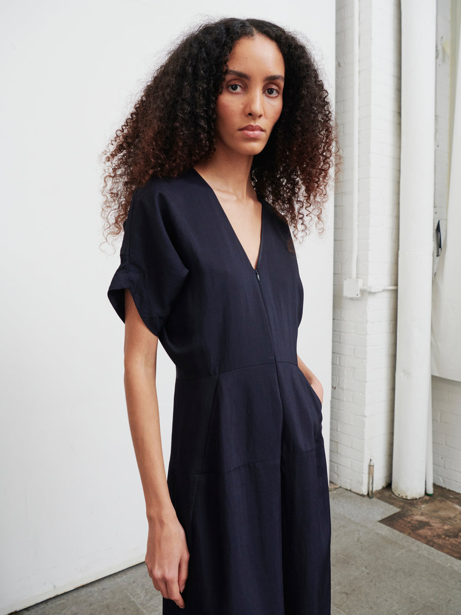 A person with long, curly hair is wearing the Aissa Eve Jumpsuit from Zero + Maria Cornejo in a minimalist room with white walls and a concrete floor. They are looking directly at the camera.