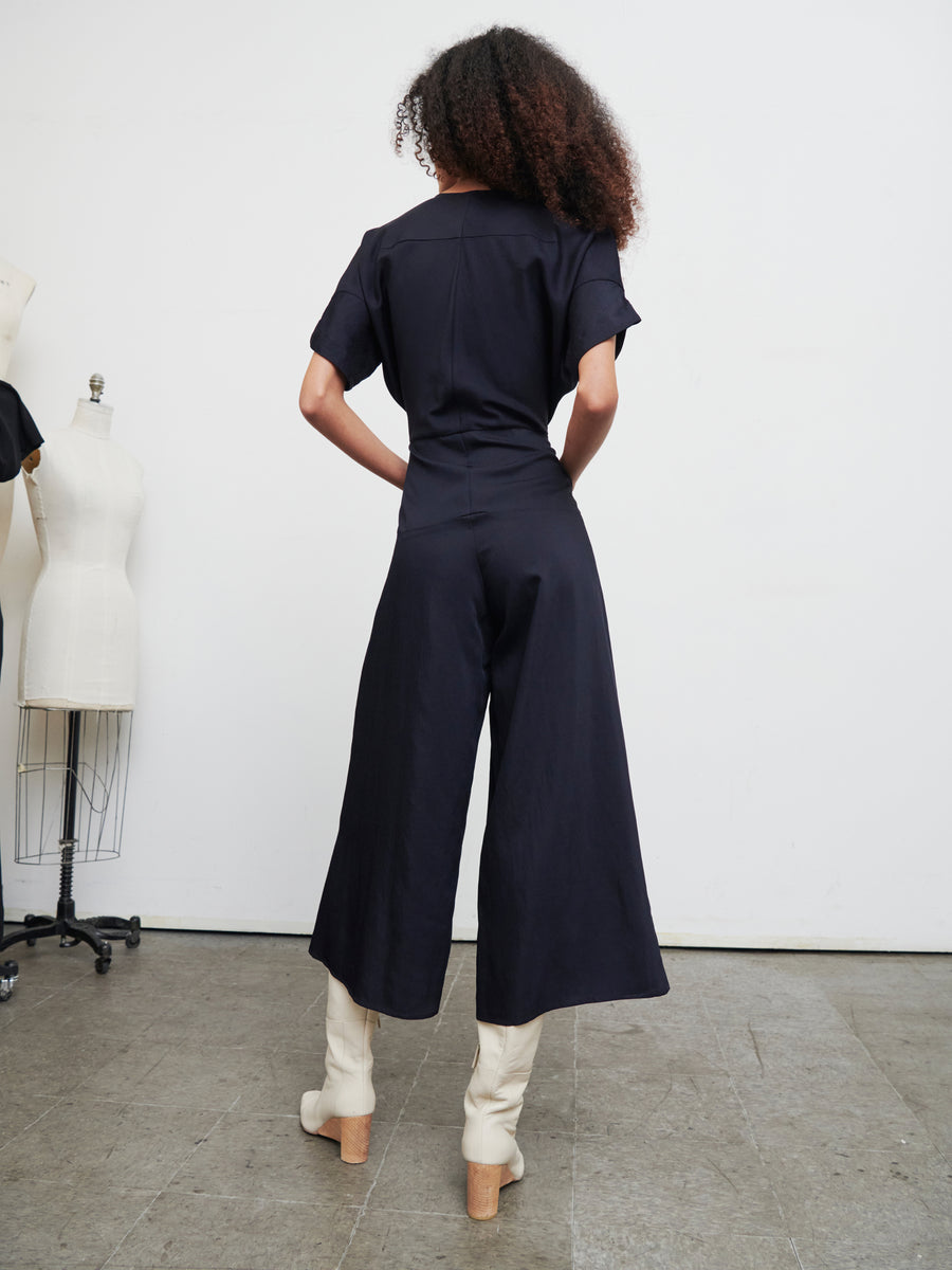 A person with curly hair stands with their back to the camera in a room filled with mannequins, wearing the Aissa Eve Jumpsuit by Zero + Maria Cornejo and white boots. The floor is tiled, and the walls are white.