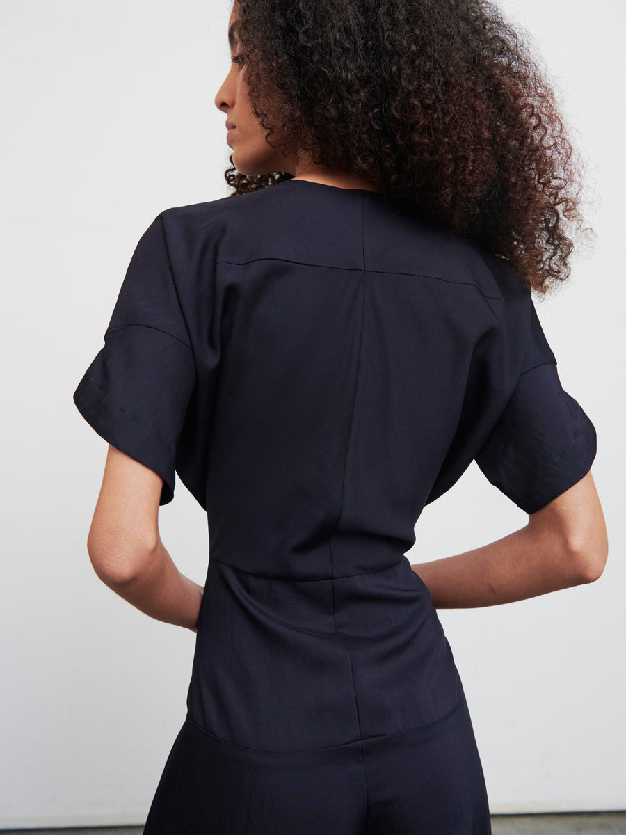 A person with curly hair stands against a white background, showcasing the back view of the Aissa Eve Jumpsuit by Zero + Maria Cornejo, a dark navy ensemble made from recycled viscose featuring short sleeves.