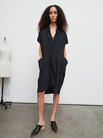 A person with curly hair stands confidently in the Fin Gaban Dress by Zero + Maria Cornejo, an asymmetrical, knee-length dress crafted from recycled viscose, paired with black slip-on shoes. Their hands rest in the dress pockets, while a dress form is visible on the left side against a plain white wall.