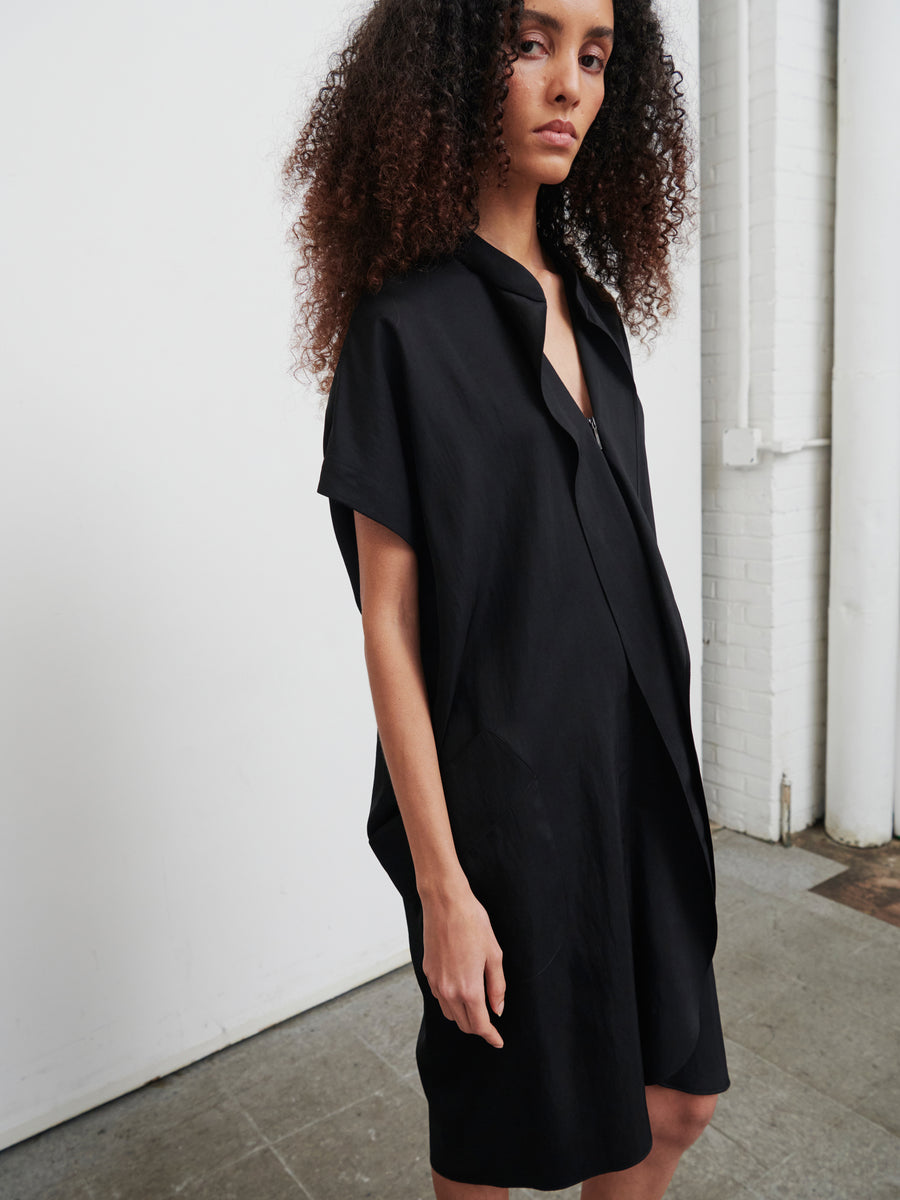 A person with curly hair stands in a minimalist interior, wearing the Fin Gaban Dress by Zero + Maria Cornejo. The dress is a loose-fitting, knee-length black piece made from recycled viscose. They have a calm expression and are positioned slightly to the side against a background of a white brick wall and gray floor.