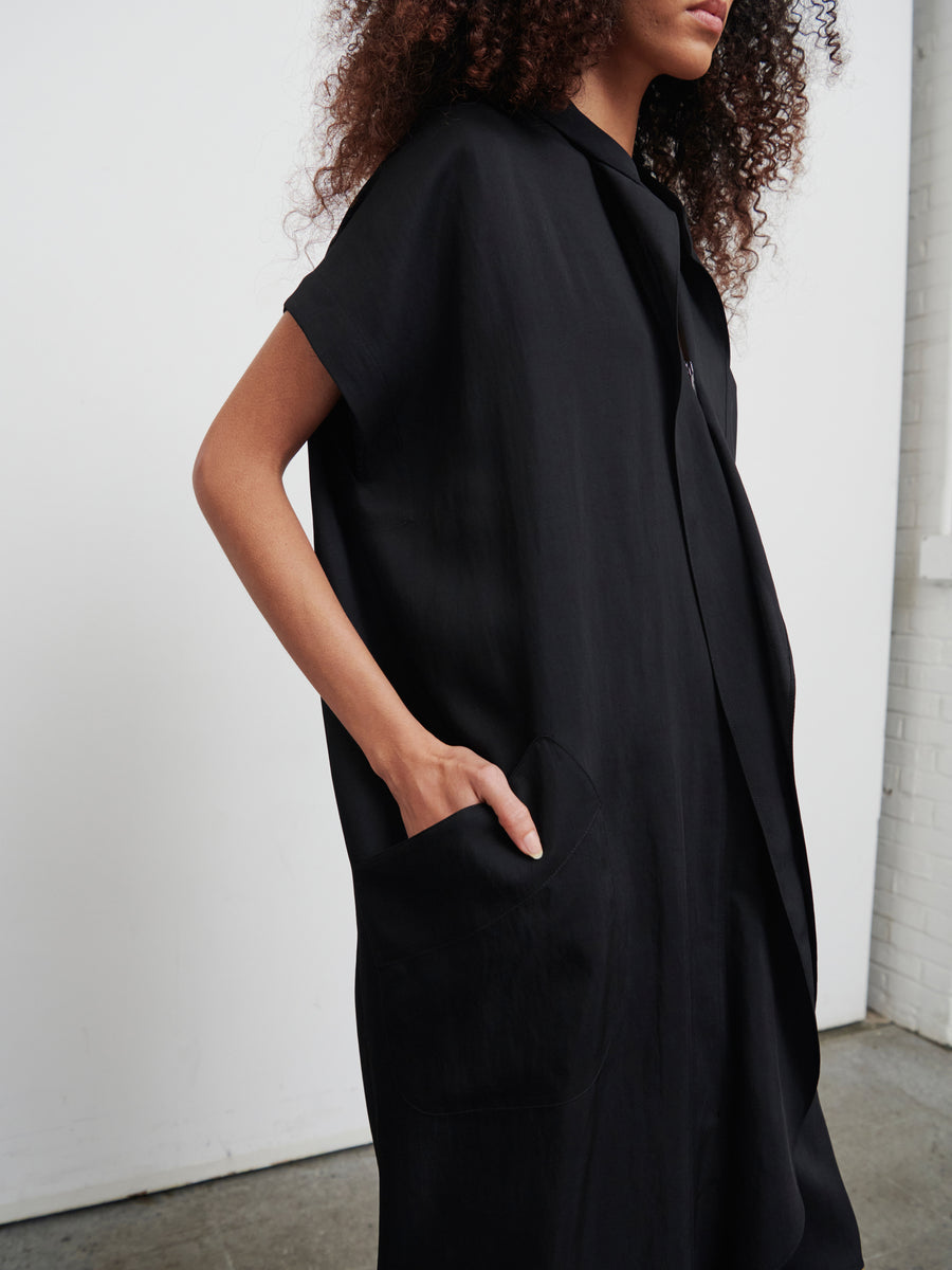 A person with curly hair wearing the loose-fitting Fin Gaban Dress from Zero + Maria Cornejo, made from recycled viscose, stands in a minimalist space. The knee-length dress features short sleeves and large pockets. Casually, the person places one hand in a pocket against the backdrop of a plain white wall and concrete floor.