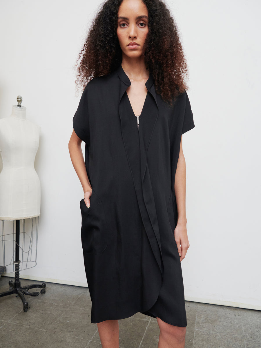 A person with curly hair stands in a room wearing the Fin Gaban Dress from Zero + Maria Cornejo, a loose-fitting knee-length black garment complete with pockets. In the background to the left, a mannequin further enhances the charm of the scene.