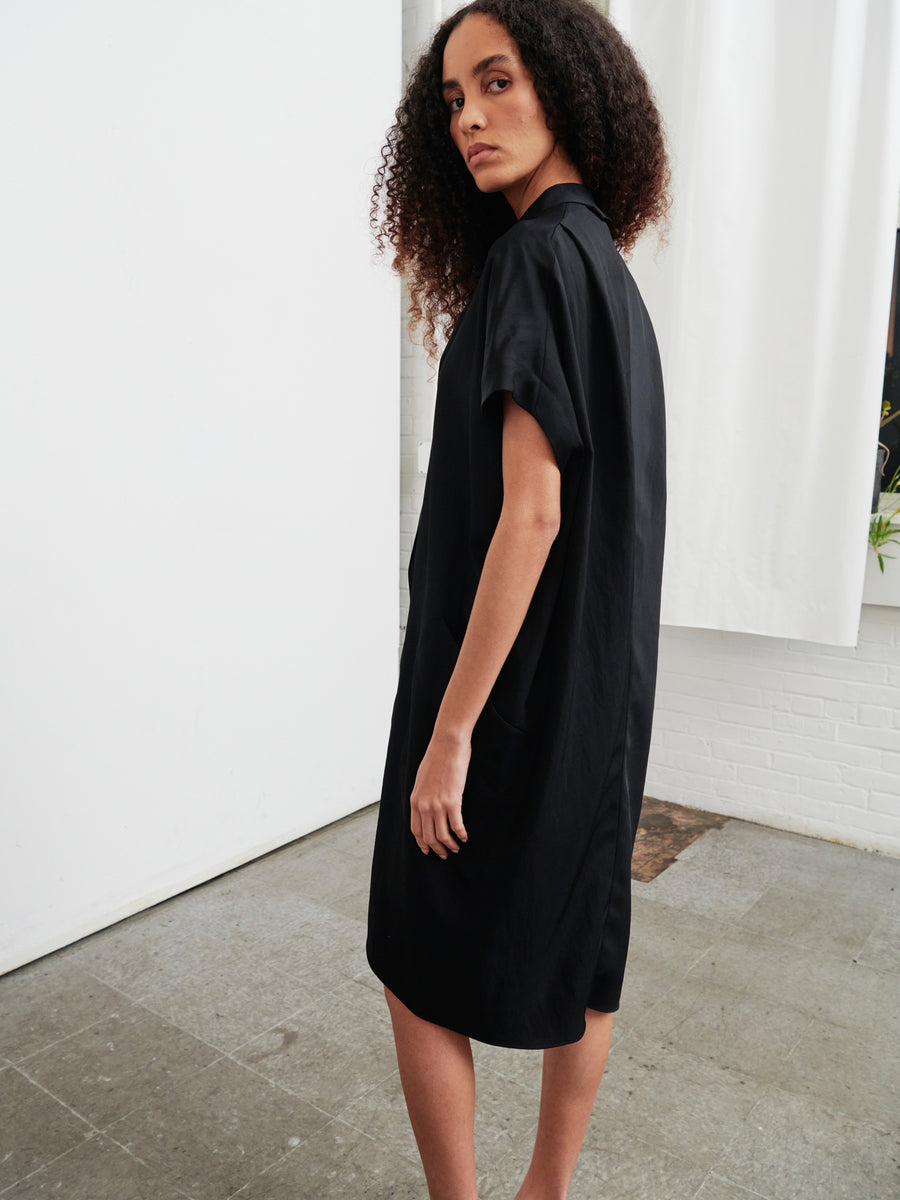 A person with long curly hair is wearing the Fin Gaban Dress by Zero + Maria Cornejo, a loose-fitting, knee-length black dress crafted from recycled viscose. They are standing on a tiled floor indoors, glancing over their shoulder, with white curtains and a brick wall in the background.