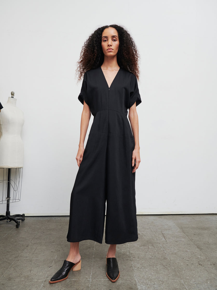 A person with long curly hair stands confidently in the Aissa Eve Jumpsuit by Zero + Maria Cornejo, featuring cropped wide legs and paired with black shoes. They are in a minimalistic room with dress forms in the background.