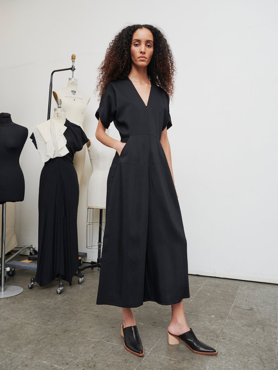 A woman with curly hair is confidently wearing the stylish Aissa Eve Jumpsuit in black, designed by Zero + Maria Cornejo, paired with black shoes. In the background, dress forms and mannequins showcase various garments in a studio setting.