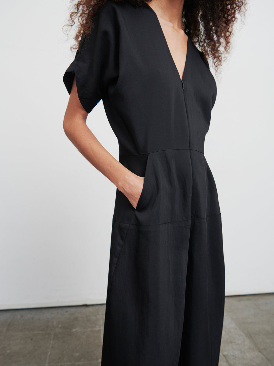 The individual is dressed in the Aissa Eve Jumpsuit by Zero + Maria Cornejo, featuring a black hue and short sleeves. They are set against a simple white backdrop, with their right hand casually placed in a pocket. The recycled viscose material of the jumpsuit looks smooth and subtly structured, showcasing cropped wide legs for a relaxed yet fashionable vibe.