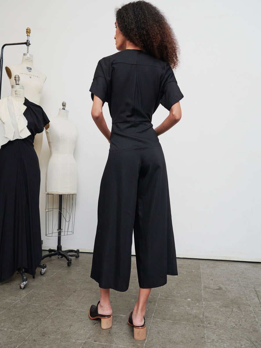 A person with curly hair stands with their back to the camera, dressed in the Aissa Eve Jumpsuit from Zero + Maria Cornejo and platform shoes. In the background, dress forms are adorned with draped fabric in a studio setting.