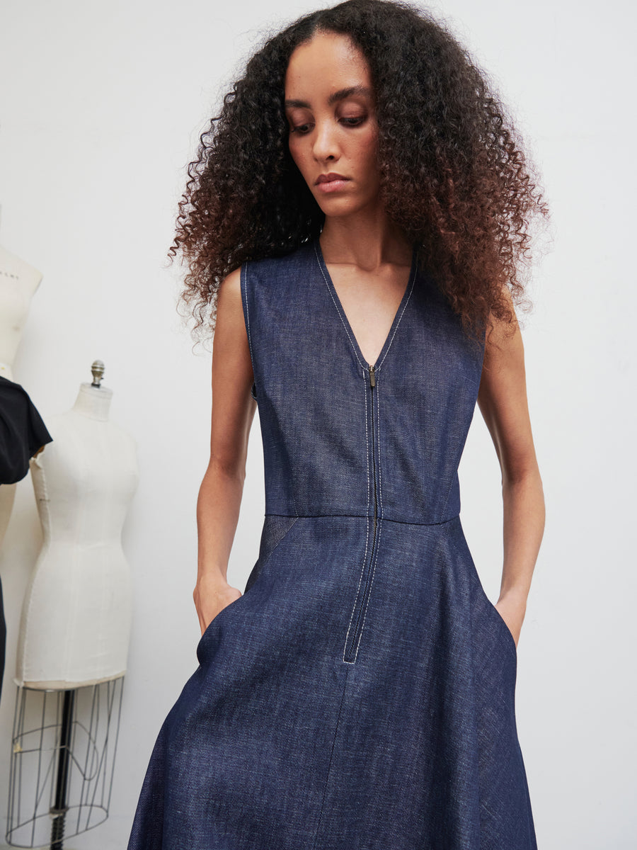 A woman with curly hair poses against a white background, wearing the Denim Wave Dress by Zero + Maria Cornejo. The dress is sleeveless with a v-neck, made from organic denim, and features a front zipper. She stands with her hands in the dress's pockets, while two dress forms are visible in the background.