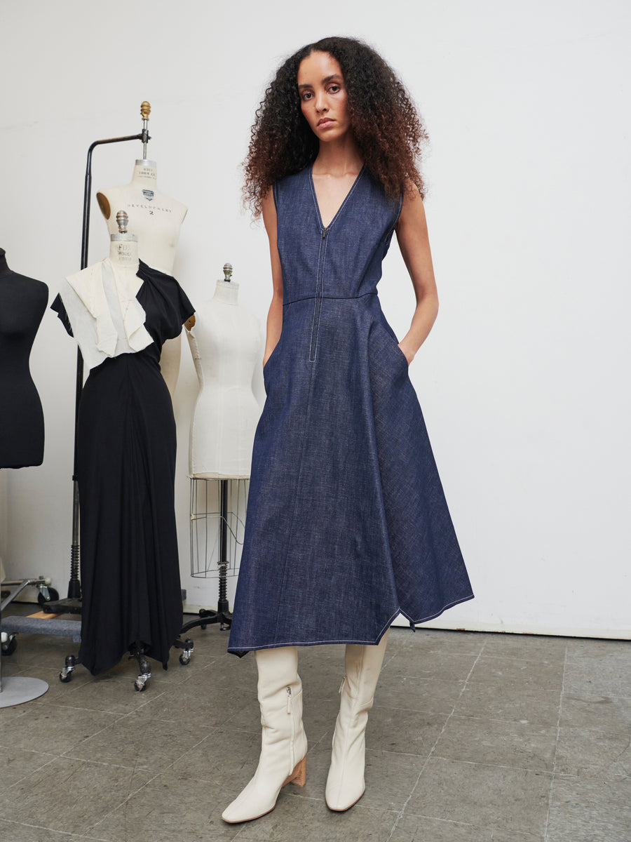 A person with curly hair is wearing the sleeveless Denim Wave Dress from Zero + Maria Cornejo paired with white knee-high boots. They stand in a fashion studio surrounded by dress forms, including a striking black gown adorned with white ruffles.