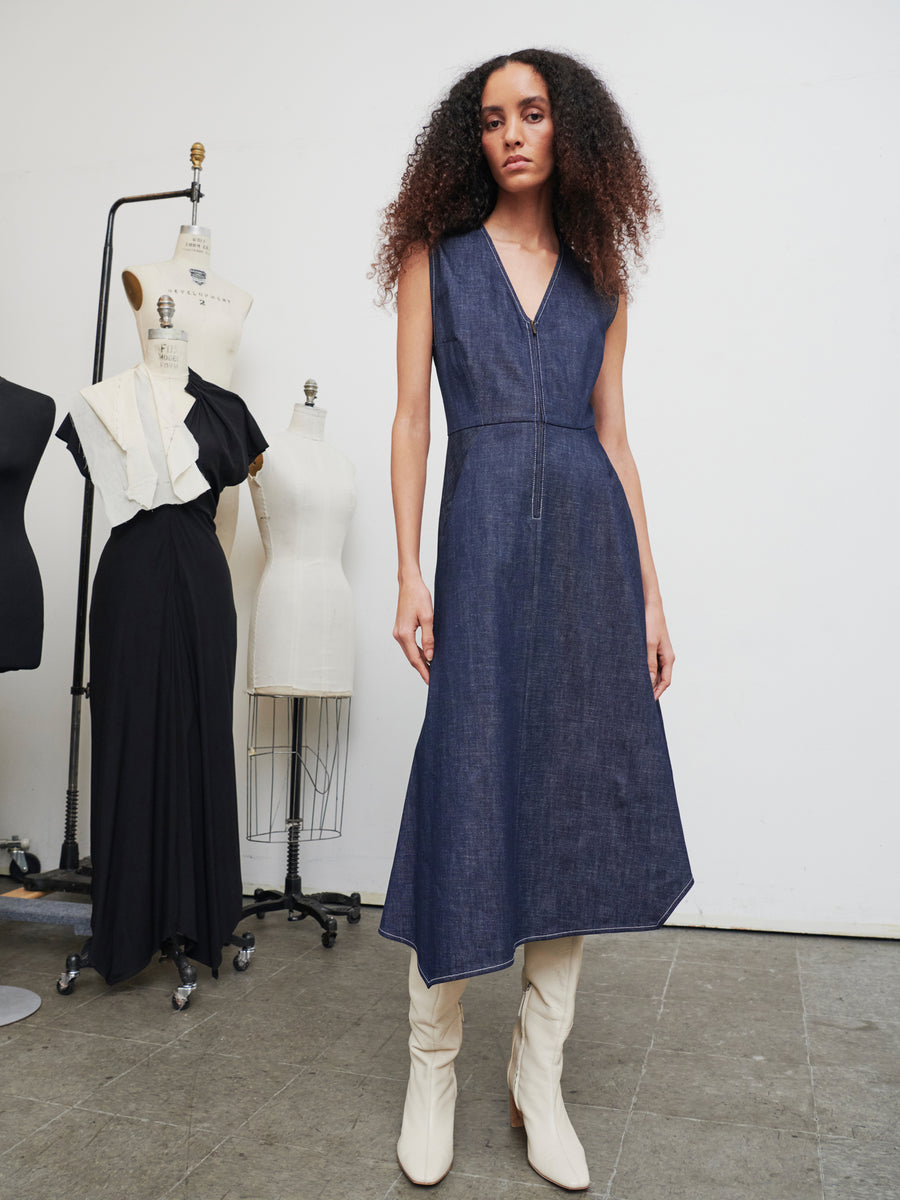 A person with curly hair wears the Denim Wave Dress from Zero + Maria Cornejo and white boots in a studio setting. Behind them are dress forms, one of which is adorned in a black dress with white accents. The room's plain gray and white walls and floor establish a minimalist backdrop.