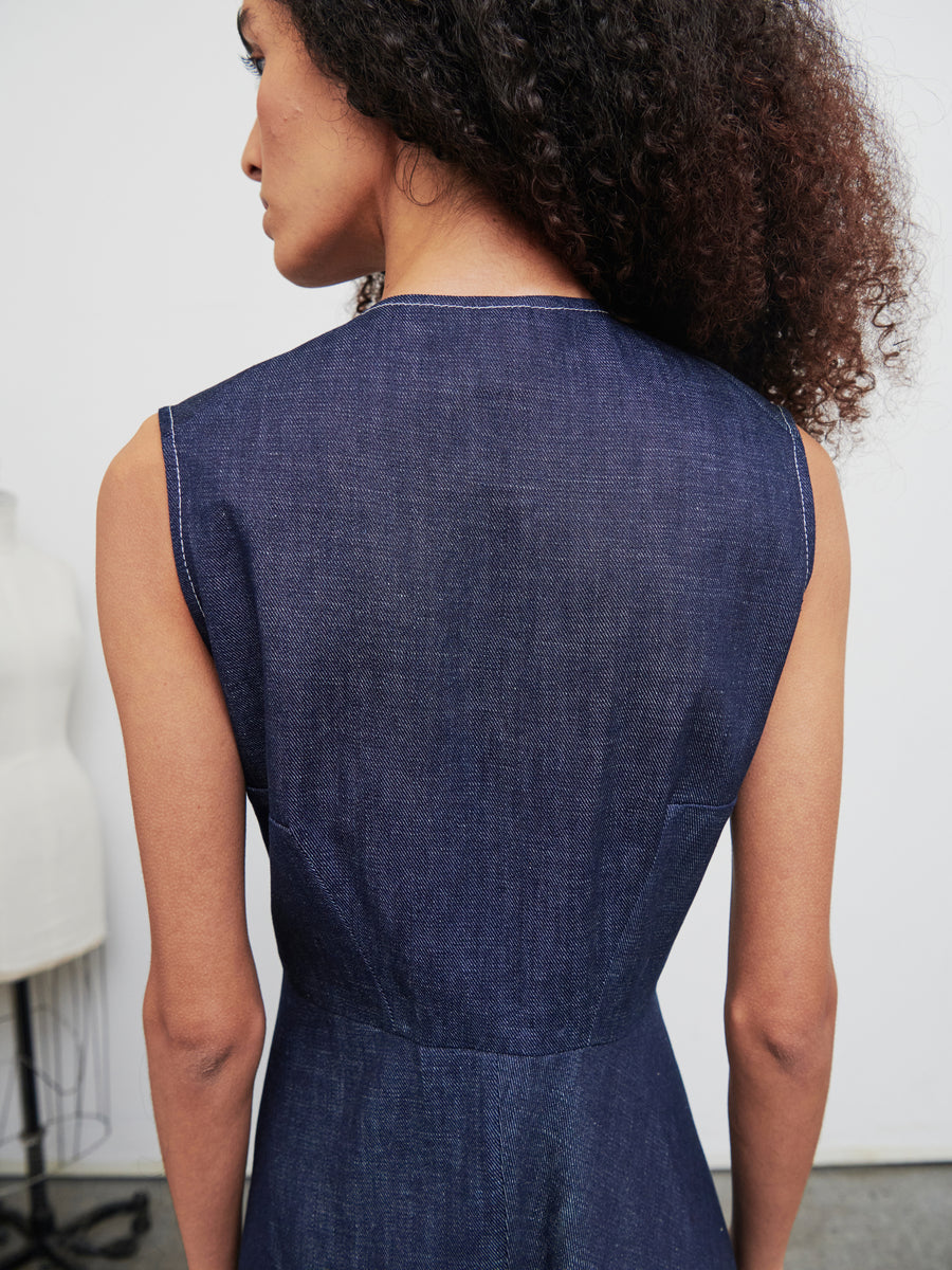 A person with long curly hair is shown from behind wearing the Denim Wave Dress by Zero + Maria Cornejo, featuring a sleeveless design in dark blue organic denim. In the blurred background to the left, there is a dress form.