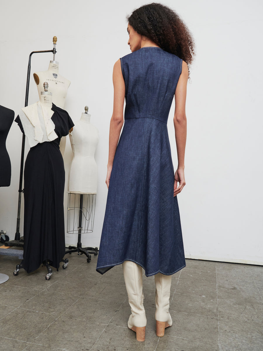 A woman with curly hair is wearing the Denim Wave Dress by Zero + Maria Cornejo along with light-colored knee-high boots. She stands in a room decorated with dress forms and other outfits on hangers, giving the impression of a fashion studio.