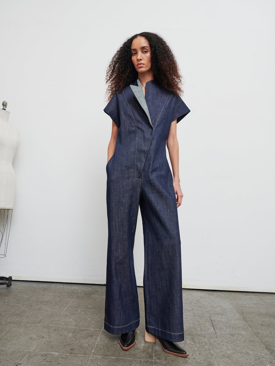 In a minimalist room, a person models the Edi Boiler Suit by Zero + Maria Cornejo, featuring wide legs and short sleeves. Their curly hair frames their face as they look at the camera, while a mannequin on the left hints at sustainable choices like GOTS-certified cotton pieces.