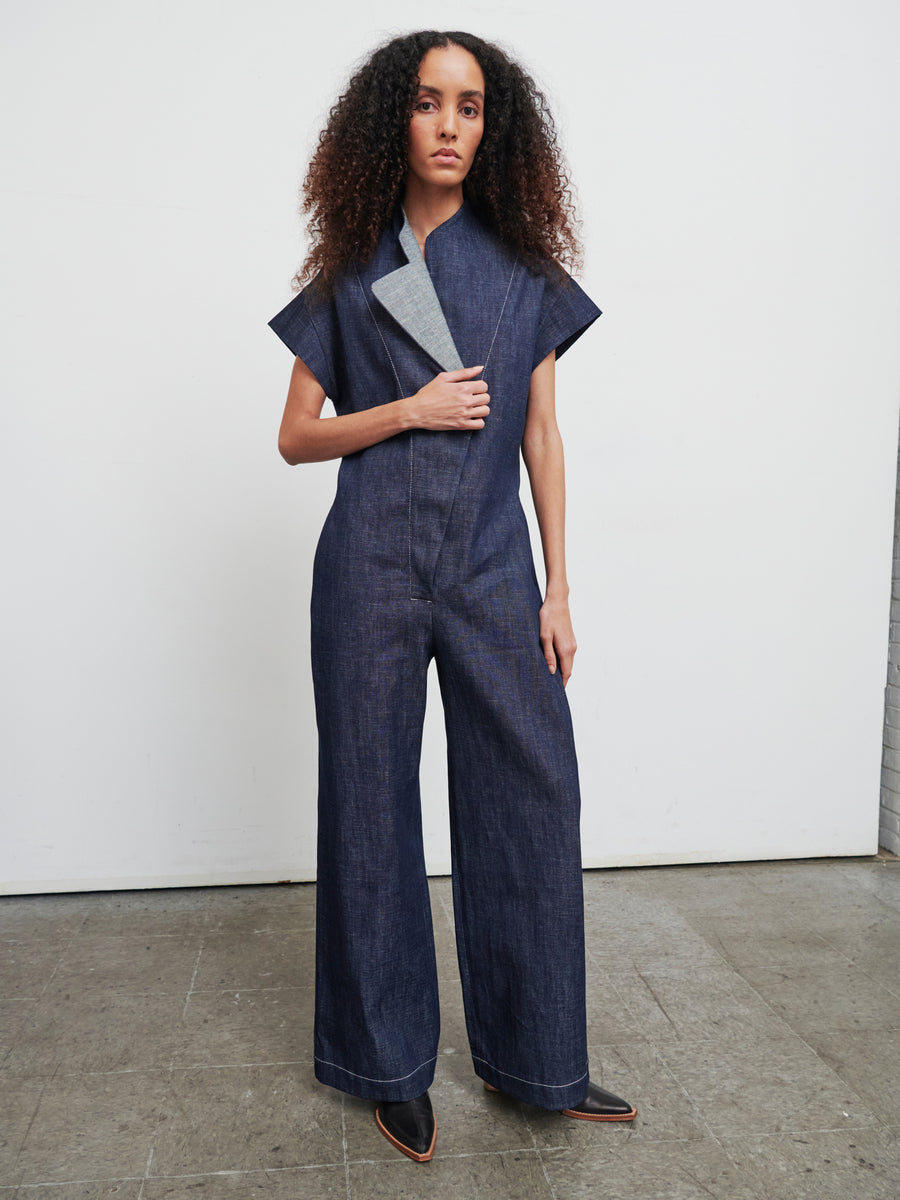 With long curly hair, a person wears the Edi Boiler Suit by Zero + Maria Cornejo, featuring wide legs and cap sleeves. They pair it with black pointed shoes, striking a confident pose on a wooden floor against a plain white wall.