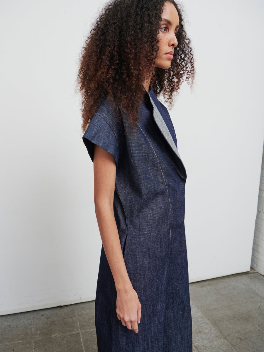 Curly-haired model in profile showcases the Edi Boiler Suit by Zero + Maria Cornejo. Standing against a white wall and concrete floor, the organic denim jumpsuit's texture and eco-friendly GOTS-certified cotton are highlighted.