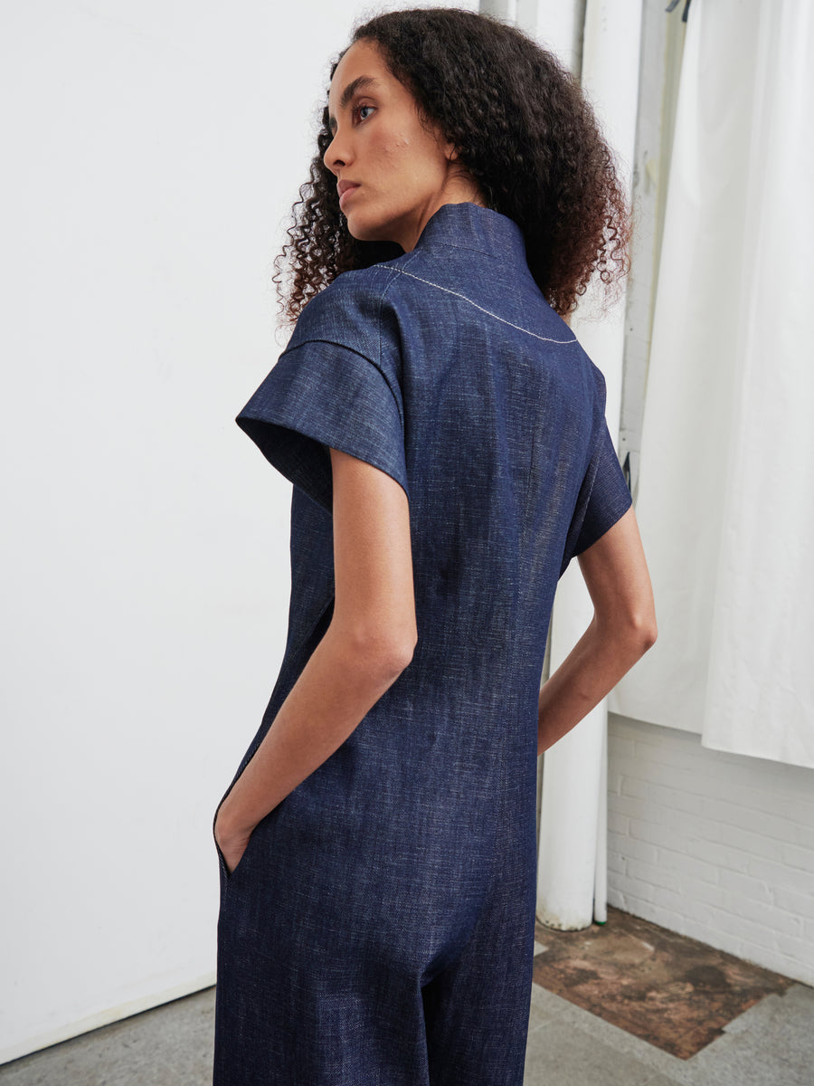 A curly-haired individual, dressed in a dark blue Edi Boiler Suit from Zero + Maria Cornejo, stands with hands in pockets, gazing to the side against a white wall with a curtain.