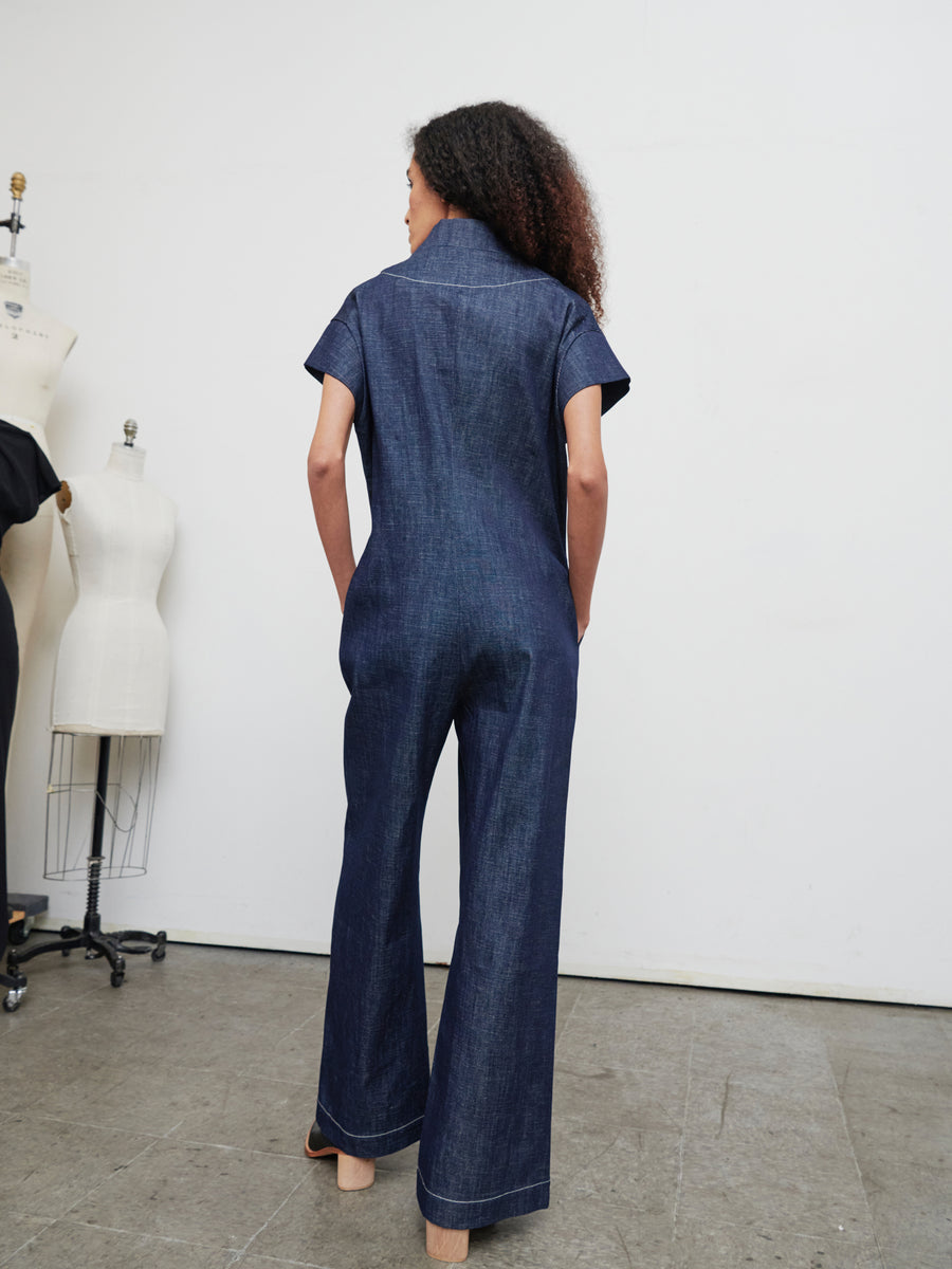 A person with curly hair stands with their back to the camera, wearing the Edi Boiler Suit from Zero + Maria Cornejo, made of GOTS-certified organic denim. They're in a studio setting with dress forms nearby.