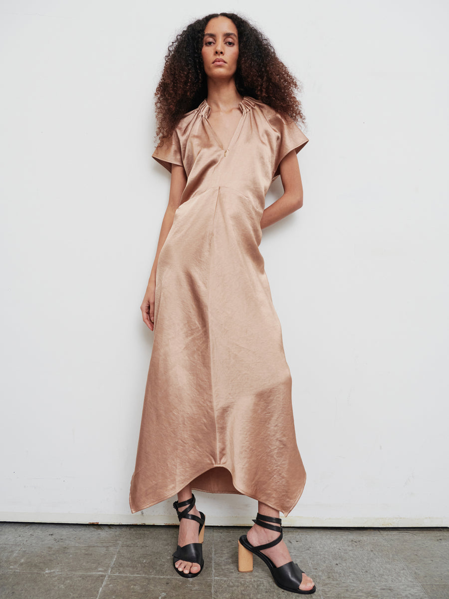 A person with curly hair wearing the Long Ruched Stella Dress by Zero + Maria Cornejo, a flowing, silky beige gown featuring gathered detailing, along with black high-heeled sandals stands against a plain white wall, looking confidently at the camera.