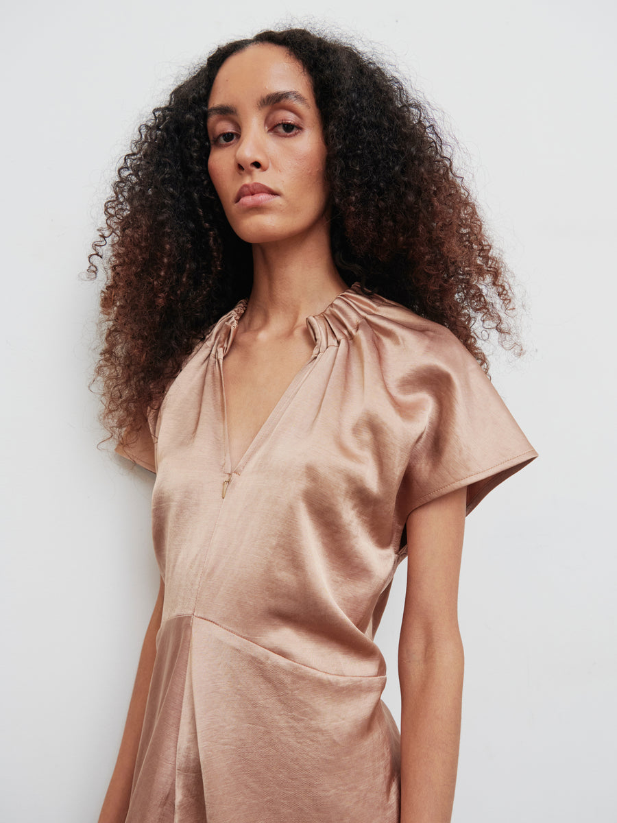 A person with curly hair wears the Long Ruched Stella Dress by Zero + Maria Cornejo. This silky, copper-colored dress features a V-neck, short sleeves, gathered detailing, and a front zip closure. The plain white background accentuates the dress's texture and sheen.