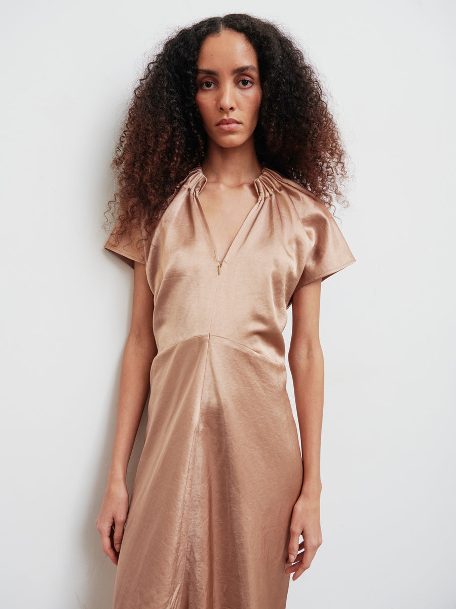 A person with long curly hair poses against a plain white backdrop, wearing the Long Ruched Stella Dress by Zero + Maria Cornejo. The dress is shiny copper-colored with gathered detailing and a subtle front zip closure, featuring short sleeves and a V-neckline. Their expression is neutral, hands resting calmly by their sides.