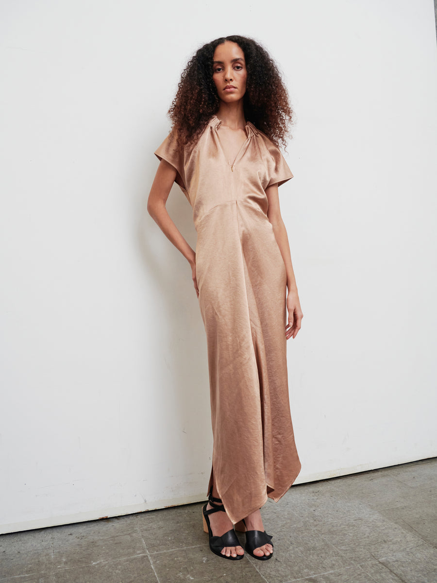 A person with curly hair stands against a white wall, wearing the Long Ruched Stella Dress by Zero + Maria Cornejo, a beige satin dress with gathered detailing and short sleeves, paired with black strappy sandals.