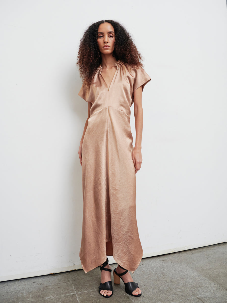 A woman with curly hair wearing the Long Ruched Stella Dress by Zero + Maria Cornejo, styled with gathered detailing and complemented by black sandals, stands against a plain white wall with a neutral expression.