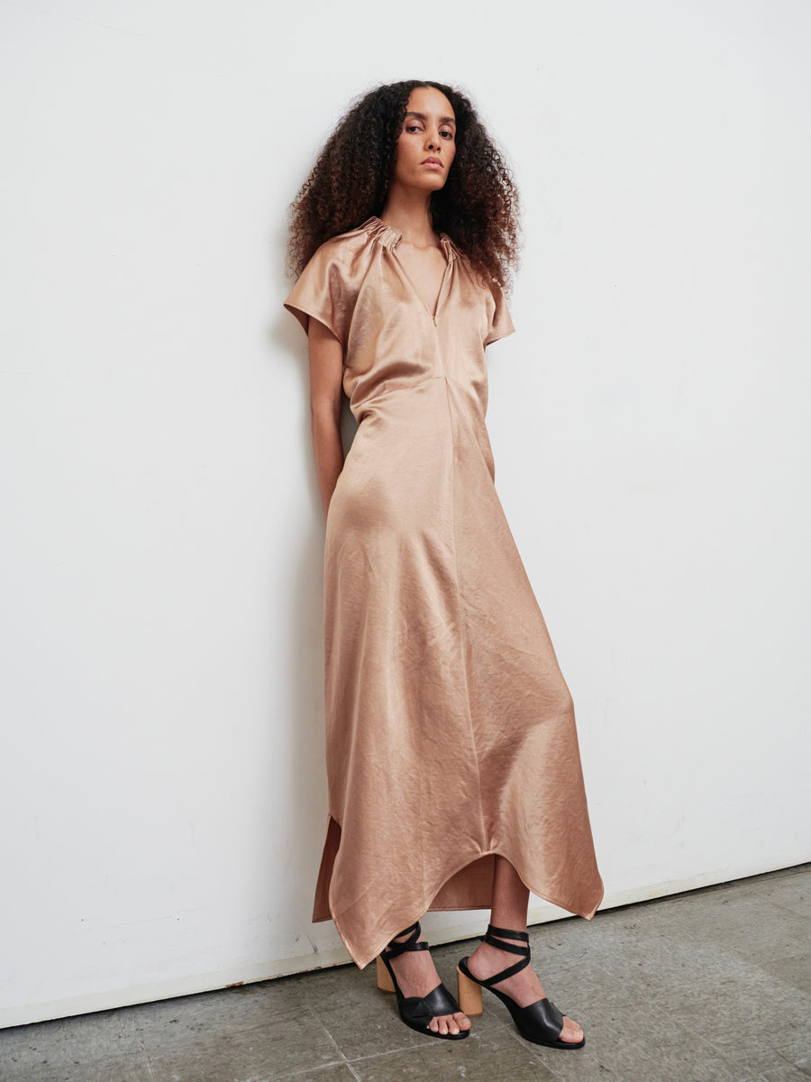 Against a white wall, a woman with long, curly hair confidently showcases the sophisticated Long Ruched Stella Dress by Zero + Maria Cornejo. Her ensemble, complete with black sandals featuring wooden heels and a modern front zip closure, exudes both elegance and poise.