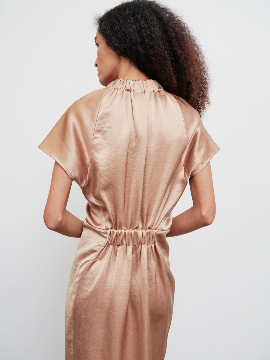 A woman with curly hair is wearing the Long Ruched Stella Dress, a shiny, short-sleeved bronze piece by Zero + Maria Cornejo. She stands with her back to the camera against a plain white background.