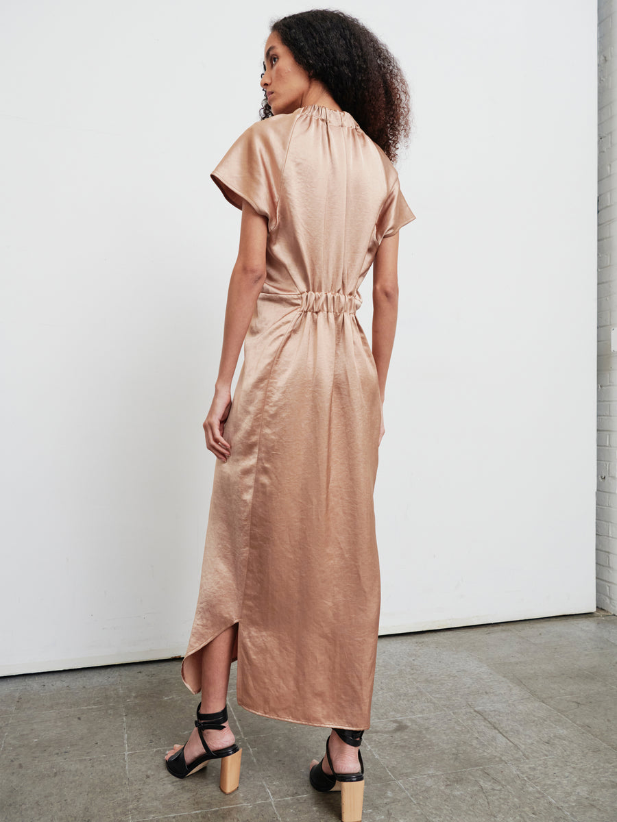 A person with long curly hair is wearing the Long Ruched Stella Dress by Zero + Maria Cornejo, which features a shiny tan satin fabric with gathered detailing, accompanied by black-heeled sandals. They are standing in a minimalistic room with a gray floor and light-colored walls, facing away from the camera.