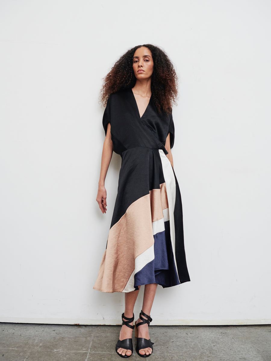 A person with curly hair stands against a white wall wearing the Patchwork Aki Wave Dress by Zero + Maria Cornejo, featuring a wrap-effect V-neck and an asymmetrical, flowing skirt with beige, white, and navy accents. They are also wearing black strappy heels.