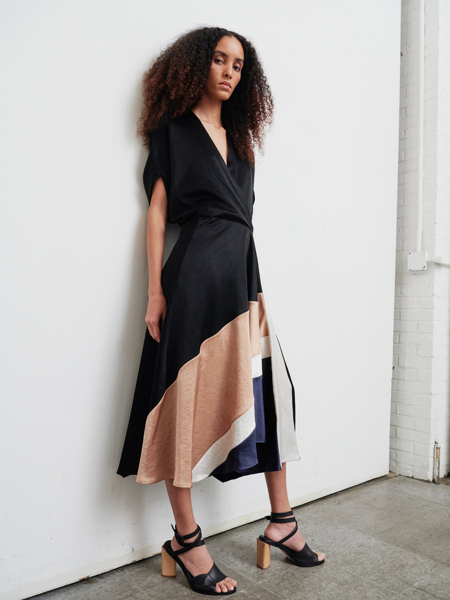 Against a white wall, a person with curly hair showcases the Patchwork Aki Wave Dress by Zero + Maria Cornejo, featuring sophisticated black, beige, and cream color blocks. They pair it with black strappy heels. The light gray floor creates a striking contrast with the pristine white brick wall.