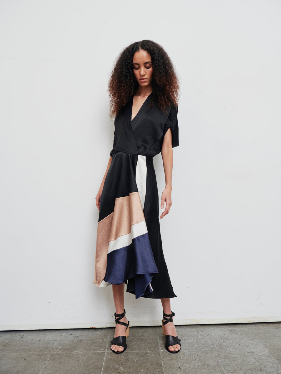 Against a plain white backdrop, a woman is seen wearing the Patchwork Aki Wave Dress by Zero + Maria Cornejo. The dress is black with asymmetrical color blocks of beige, white, and blue, styled in a wrap-effect v-neck fashion. Her curly hair frames her face as she gazes downward in a relaxed posture, complemented by black strappy heels.