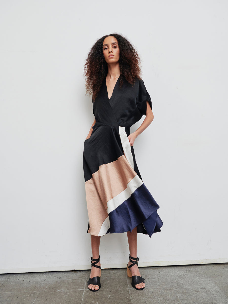 A woman stands against a white wall wearing the chic Patchwork Aki Wave Dress by Zero + Maria Cornejo. This linen blend dress features a wrap-effect V-neck and is adorned with tan, white, and navy color blocks at the hem. Her long curly hair cascades down as she strikes a pose in black strappy heels, hands casually tucked into her dress pockets.