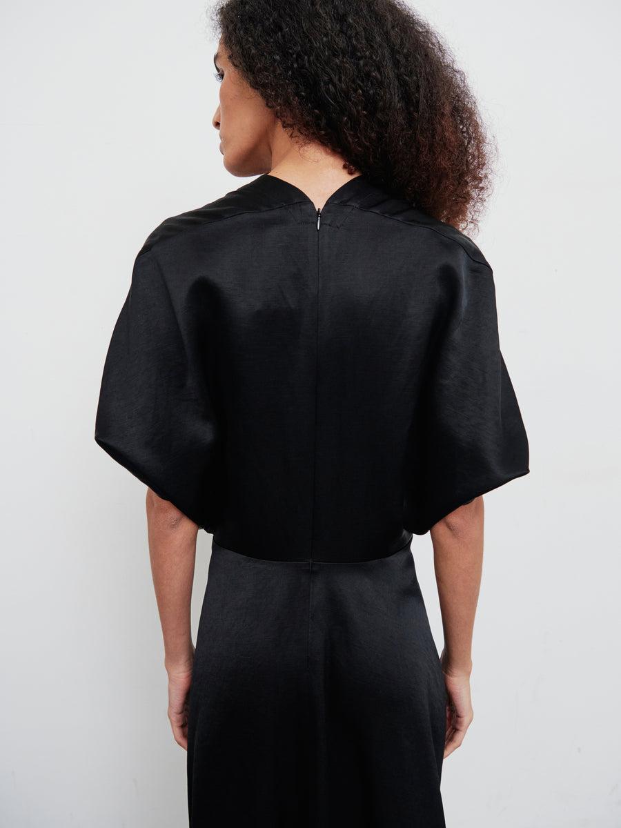 A person with curly hair is standing with their back turned, wearing the Patchwork Aki Wave Dress by Zero + Maria Cornejo, which features wide short sleeves and an asymmetrical skirt. The background is plain white.